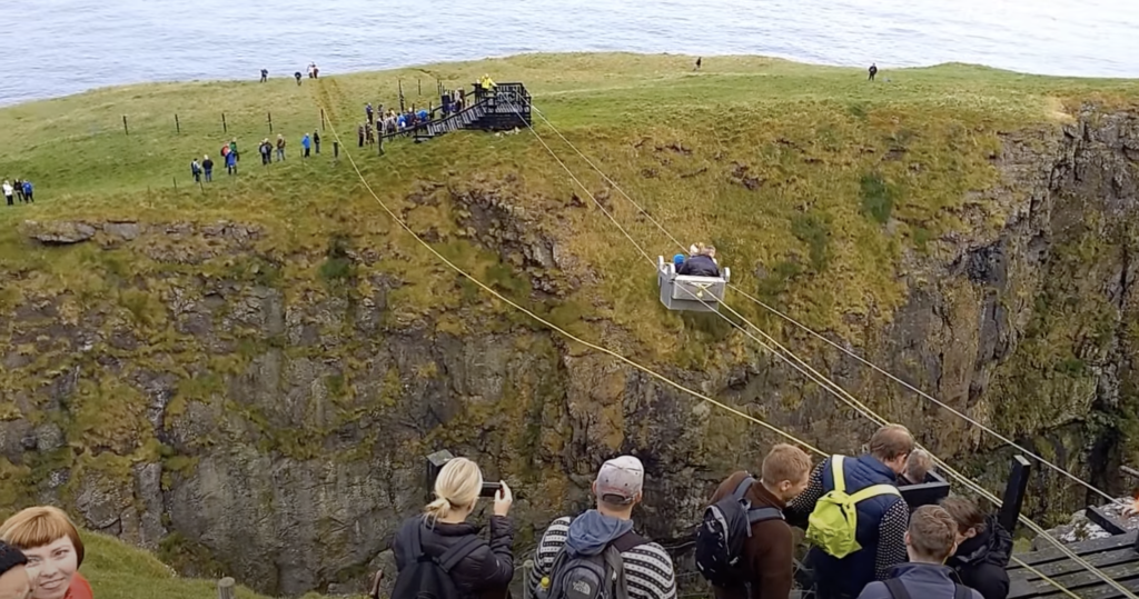 An aerial cable car car crossing between the island of Steymoy and the sea stack called Stakkurin. People are waiting for their turn on both sides of the expanse.